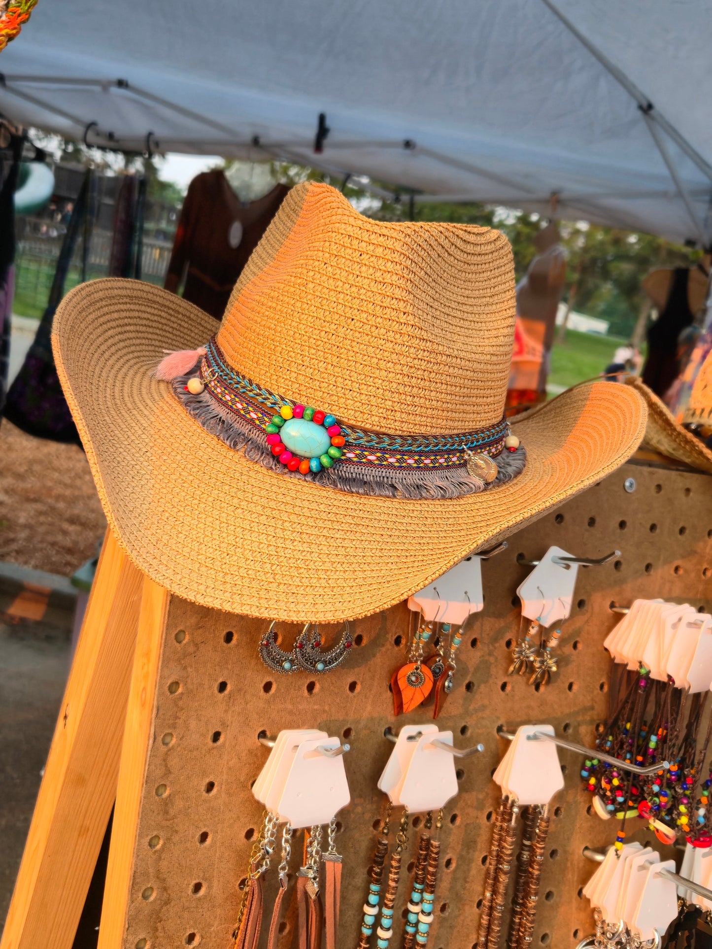 Straw boho cowboy hat with decorative band on a pegboard with jewelry at an outdoor market.