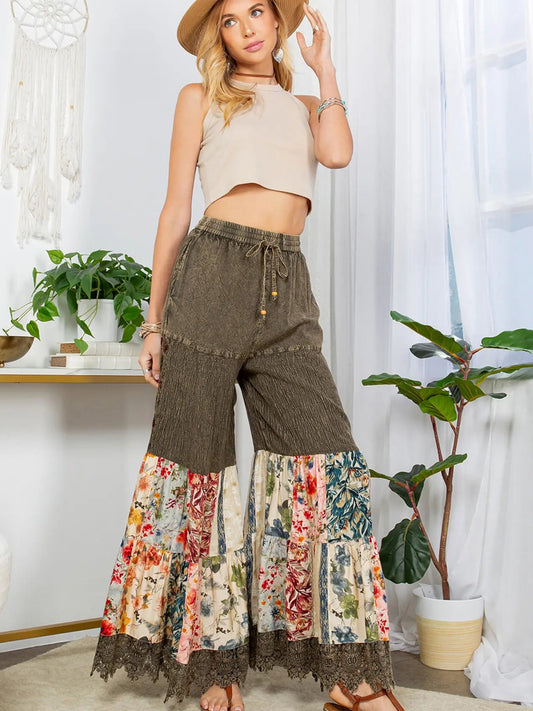 Woman wearing a beige sleeveless top and brown wide-leg pants with floral patchwork, standing in a room with plants and decor.